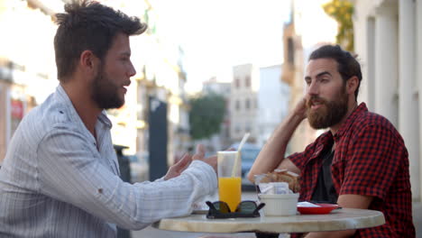 Dos-Amigos-Varones-Hablando-En-Una-Mesa-Fuera-De-Una-Cafetería,-Ibiza